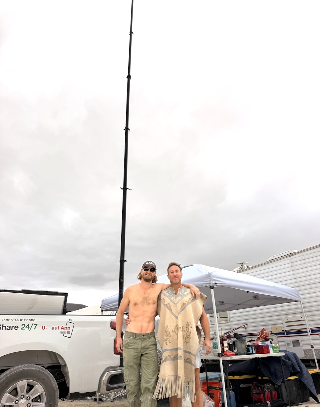 Two people standing in front of the fully extended telescopic mast at camp, the mast rising well above a pickup truck, shade canopy and RV under an overcast playa sky