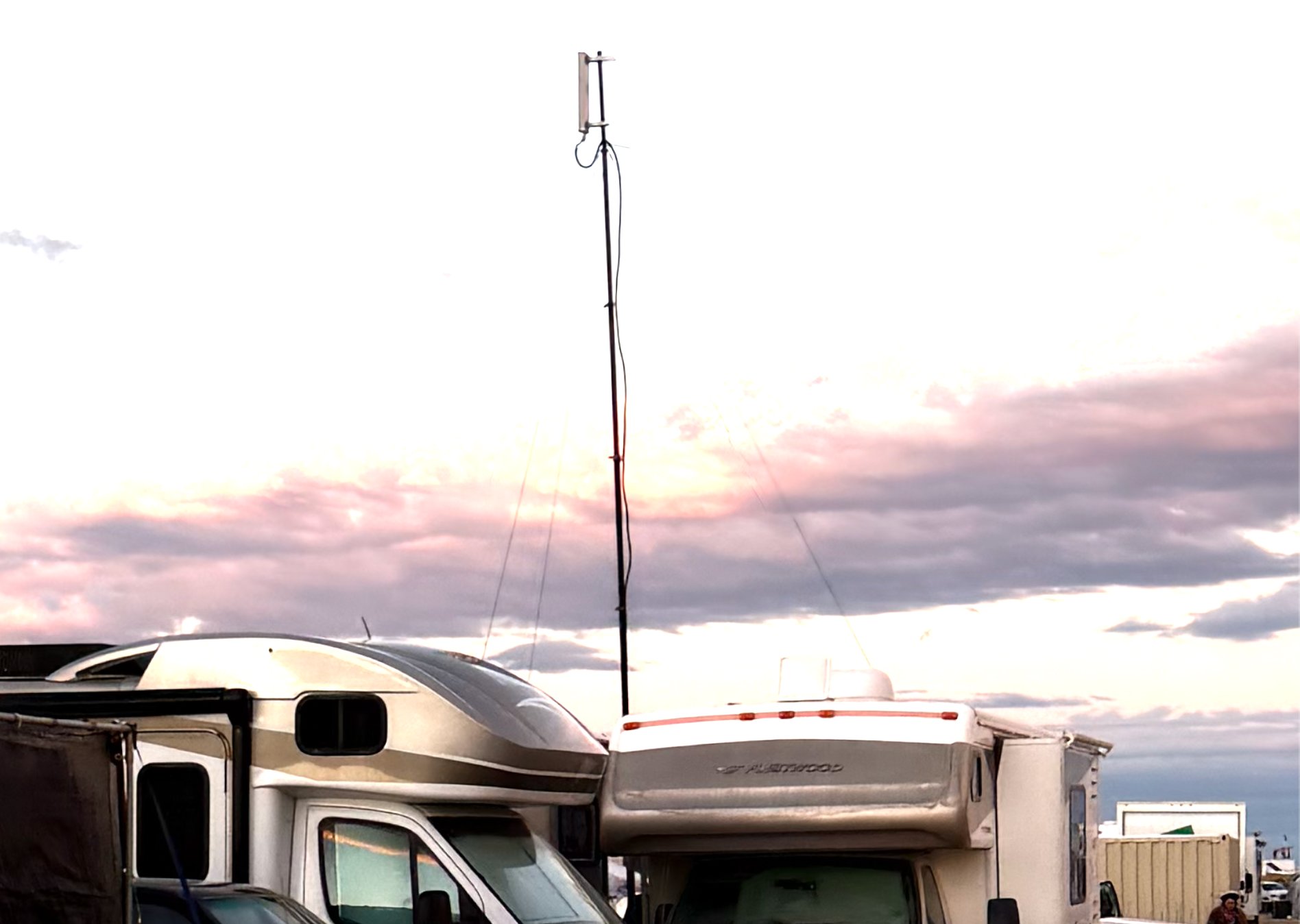 Guyed telescopic mast carrying a sector antenna, rising above parked RVs against a pink dusk sky on the open playa